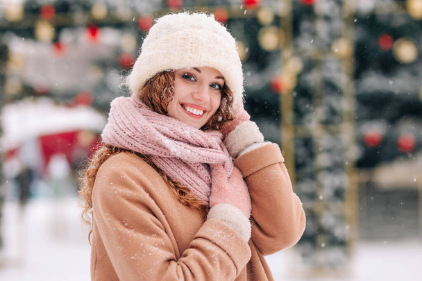 Une fille bouclée aux cheveux roux tient son chapeau avec ses mains. Joyeux sourire femme dans un manteau posant sur le fond d'un arbre de Noël décoré de la ville par une journée d'hiver enneigée - Photo, image