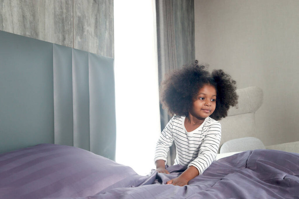 Cute little African girl child with black curly hair making a bed after waking up in morning at bedroom, kid help parents to do housework. Responsible training for children - Zdjęcie, obraz