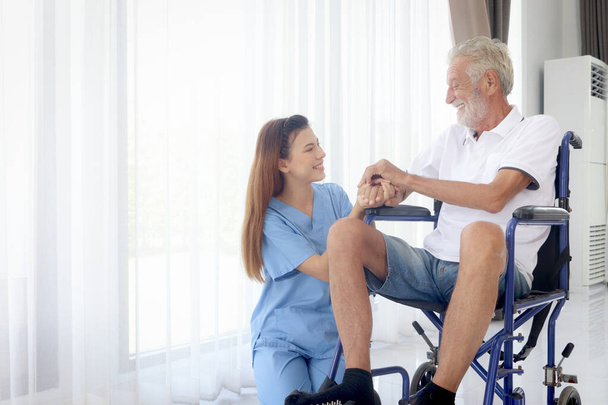 Happy senior patient in wheelchair with beautiful female nurse taking care of him at home, disabled elderly man sits in wheelchair, woman doctor helping mature grandfather, nursing medical health care - Photo, Image