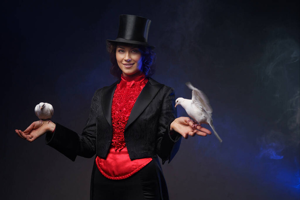 Веселая колдунья, одетая в волшебный костюм и черную цилиндр, совершает очаровательные трюки с белыми голубями на темном фоне, освещенном синим светом. - Фото, изображение