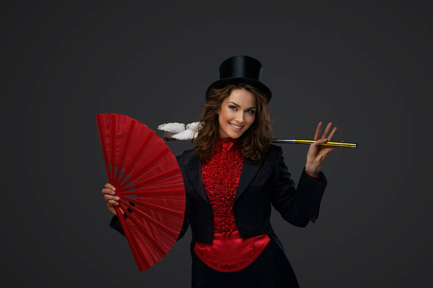 Joyeux illusionniste féminin effectuant des tours de magie avec des colombes blanches tout en tenant un grand ventilateur rouge sur un fond gris. - Photo, image
