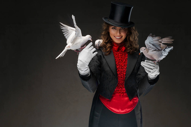 Een portret van illusionist in een magiër kostuum en zwarte hoed die goocheltrucs uithaalt met witte duiven tegen een donkere achtergrond - Foto, afbeelding