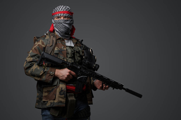 Soldato fanatico radicale mediorientale che indossa un keffiyeh bianco e uniforme camuffata da campo, armato con un fucile automatico, su sfondo grigio - Foto, immagini