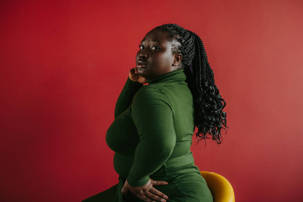 Beautiful curvy African woman with braided hair looking at camera while sitting against red background - Фото, зображення