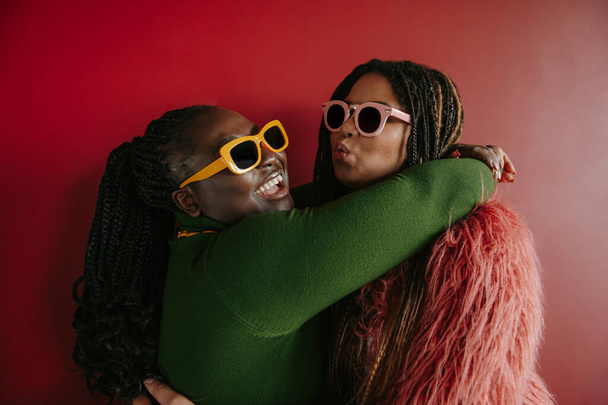 Two joyful plus size African woman with braided hair embracing on red background together - Foto, afbeelding