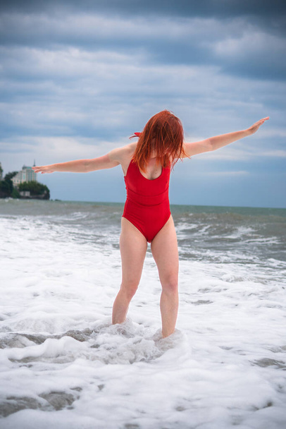 赤い閉じられた水着の幸せな女性は,波で楽しんでいます. ストーミー海,雲。 ロッキービーチ。 縦の写真。 水泳,冷たい水,大きな危険な波のための悪天候 - 写真・画像
