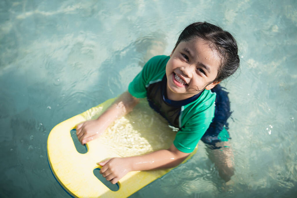 Asian child kid girl smiling funny wearing swimming suit using kick board to swimming or leaning training on swimming pool in water park. Water activity for children on summer holiday. - Photo, Image