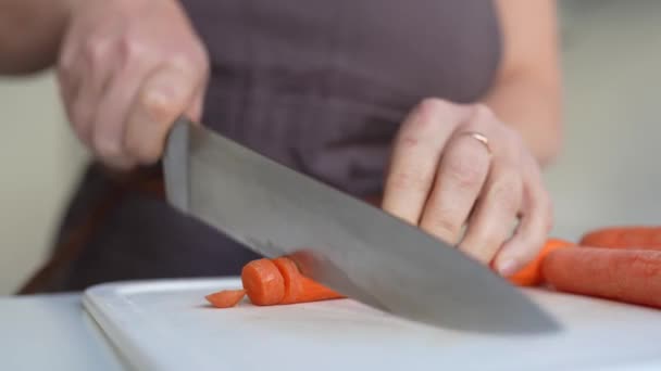 nerozpoznatelná žena v hnědé zástěře krájí mrkev s velkým kuchyňským nožem na palubě, v domácí kuchyni. - Záběry, video