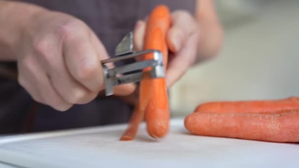 figura mulher irreconhecível em avental marrom descascando cenouras com faca de descascar na cozinha - Filmagem, Vídeo