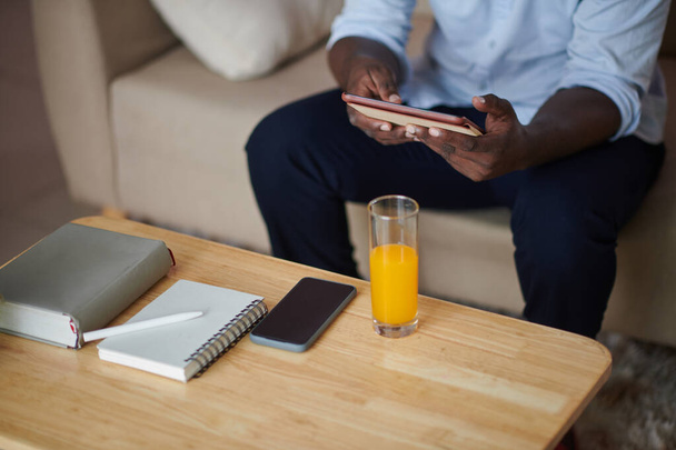 Zugeschnittenes Bild eines Geschäftsmannes, der mit Planer, Smartphone und einem Glas Orangensaft vor einem Couchtisch sitzt und E-Books liest - Foto, Bild