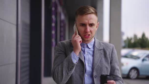 Vihainen liikemies pitelee kahvia kävellessään ulkona ja puhuessaan matkapuhelimella. Liiketoiminta, työ, teknologia. Hidastus - Materiaali, video
