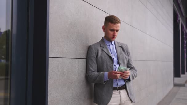 Young man counting money, standing outdoors near wall and putting it in his pocket. Business, payment concept. Slow motion - Footage, Video