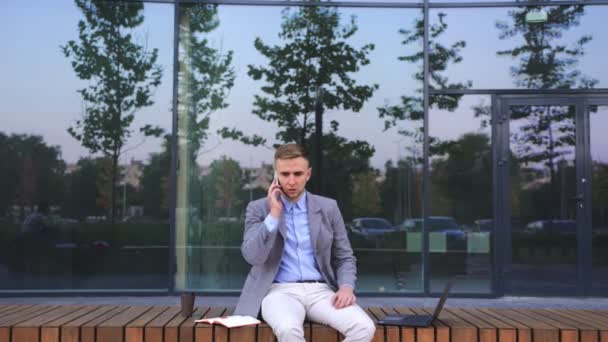 Järkyttynyt liikemies istuu penkillä puhuessaan matkapuhelimeen kaupungissa. Liiketoiminta, työ, teknologia. Hidastus - Materiaali, video