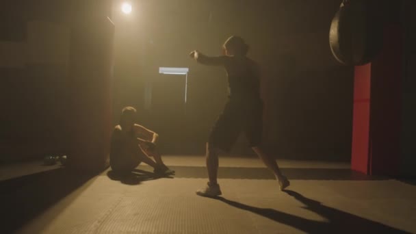 Largo tiro de deportista entrenamiento golpes durante el entrenamiento de boxeo mientras su amigo descansa en el suelo en el gimnasio débilmente iluminado - Imágenes, Vídeo