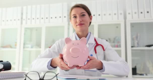 Lékařský terapeut drží prasátko v bance. Náklady na úsporu nákladů na zdravotní péči a zdravotní pojištění - Záběry, video
