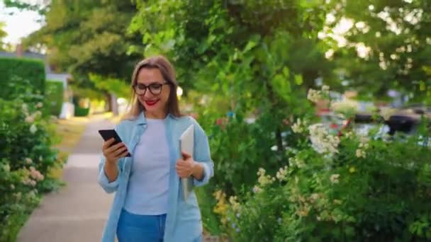 Mujer feliz con laptop y smartphone en sus manos caminando por el floreciente jardín o calle verde después del día de trabajo. - Metraje, vídeo