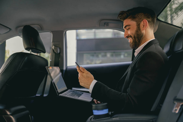 Όμορφος επιχειρηματίας με κοστούμι χρησιμοποιώντας τον προσωπικό υπολογιστή και χρησιμοποιώντας το κινητό τηλέφωνο στο ταξί - Φωτογραφία, εικόνα