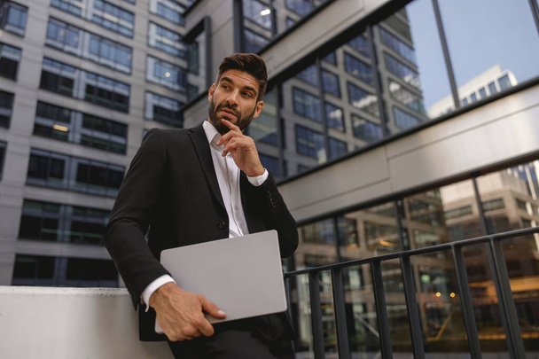 Élégant gestionnaire masculin debout avec ordinateur portable sur la terrasse du bureau et regarde ailleurs. Photo de haute qualité - Photo, image