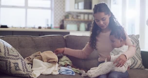.Folding laundry, mother and baby at home for bonding, chores and quality time together. Young woman multitask with an infant child, clothes and housework or cleaning on a sofa in a family lounge - Footage, Video