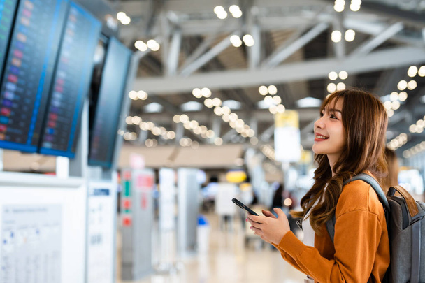 Fiatal ázsiai nő a nemzetközi repülőtéren, mobiltelefont használ és ellenőrzi a repülést a repülési információs táblán - Fotó, kép