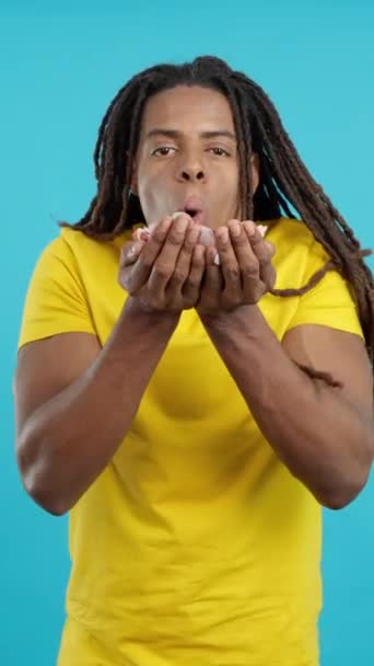 Happy latin man with dreadlocks blowing confetti in studio with blue background - Footage, Video
