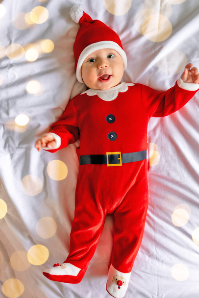 un pequeño bebé lindo en un traje de Santa acostado en la cama, un niño feliz está celebrando la Navidad y el Año Nuevo en casa - Foto, imagen