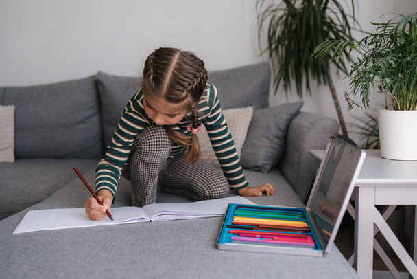 Beautiful little girl is drawing in an album while sitting on the sofa. Girl uses pencils. High quality photo - Photo, Image