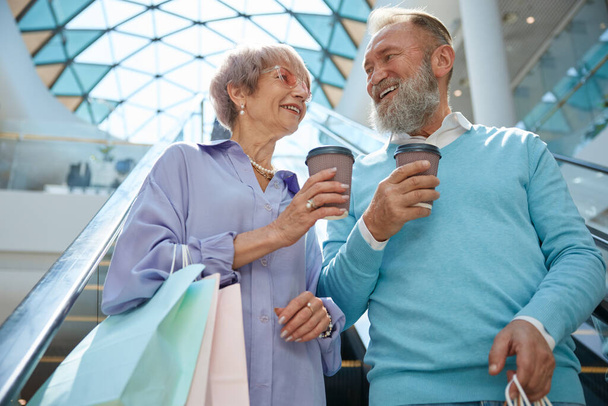 Idősebb pár, táskában és kávés sapkában vásárolnak, miközben a bevásárlóközpont mozgólépcsőjén utaznak, élvezve az együtt töltött időt. - Fotó, kép