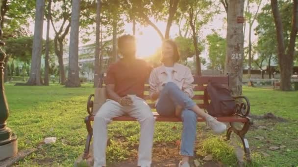 Pełne ujęcie łuku młodej pary trzymającej się filiżanek kawy i rozmawiającej siedząc razem na ławce w parku o zachodzie słońca - Materiał filmowy, wideo