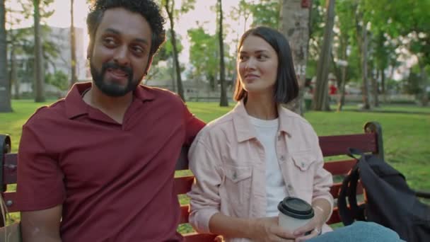 Foto de arco medio de joven pareja diversa sosteniendo tazas de café para llevar y conversando mientras descansan juntos en el banco en el parque al atardecer - Imágenes, Vídeo