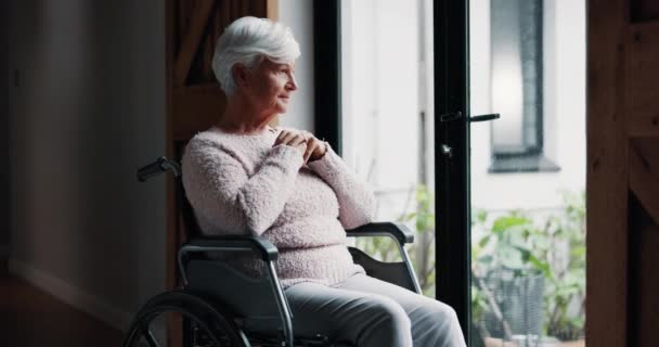 Oudere vrouw, rolstoel en denken door raam in pensionering voor hoop, droom of geloof op hoge leeftijd thuis. Eenzame oudere vrouw of persoon met een handicap in verwondering, beslissing of kijken uit in verdriet. - Video