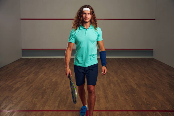 Retrato de hombre sosteniendo raqueta de squash caminando entre cancha deportiva. Jugador de racquetball seguro y deporte profesional - Foto, Imagen