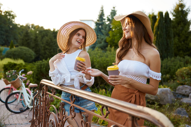 Zwei junge Modefrauen unterhalten sich stehend auf einer Brücke über See oder kleinen Fluss im Park. Schöne Natur, positive Menschen und ein glückliches Wochenende - Foto, Bild
