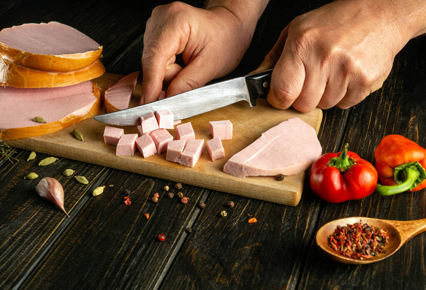 Krojenie gotowanej kiełbasy na desce do przyrządzania sałatki. Nóż w ręku kucharza na stole kuchennym - Zdjęcie, obraz