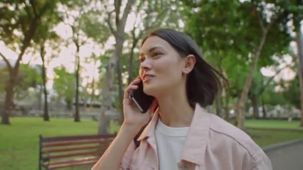 Jonge vrolijke vrouw glimlachen en chatten op mobiele telefoon tijdens het wandelen in het park bij zonsondergang - Video