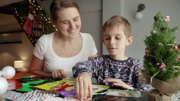 Onnellinen hymyilevä äiti katselee poikaansa tehden perinteisiä joulupostikortteja ja käsintehtyjä koristeita. - Materiaali, video
