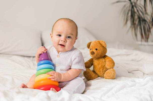 Bebê bonito brincando com pirâmide de brinquedo colorido arco-íris e ursinho de pelúcia sentado na cama no quarto ensolarado branco. Brinquedos para crianças. Criança com brinquedo educativo - Foto, Imagem