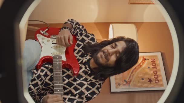 Tiro vertical medio a través de la luz selfie del joven músico de Oriente Medio sentado en casa, tocando la guitarra eléctrica roja e interpretando una canción emocional, mirando a la cámara - Imágenes, Vídeo