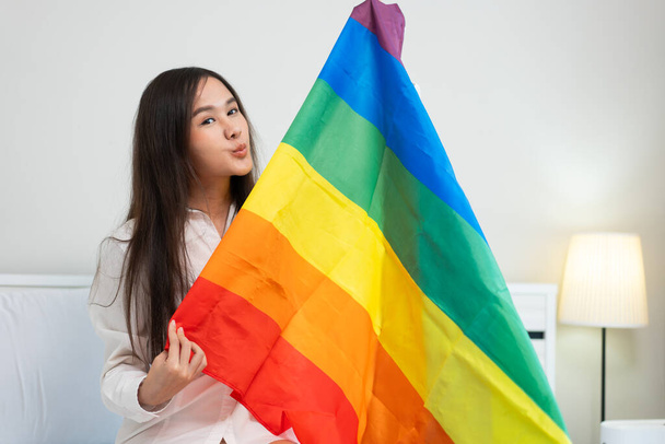 窓の横に座っているベッドの上に虹の旗を持って座っている幸せなアジアのレズビアン少女の肖像画. 関係のコンセプトで暮らす陽気な若い成人女性. LGBTQライフスタイルプライド月間 - 写真・画像