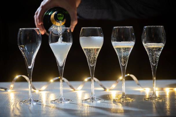 New year party, pouring of brut champagne bubbles cava or prosecco wine in tulip glasses with garland  lights on dark background - Photo, Image