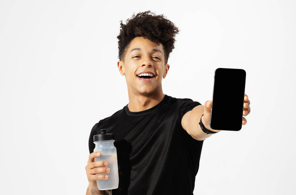 Athletic young man shows cell phone with empty mockup screen, holds gym water bottle on white studio backdrop. Sportsman advertising new sport training app, ready for workout - Foto, Imagem