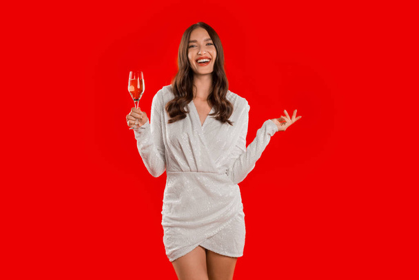 Die lächelnde Millennial Lady im glänzenden Kleid hebt ihr Champagnerglas zum festlichen Toast, ihre fröhliche Feier vor roter Studiokulisse verkörpert Urlaubsstimmung. Neujahrsparty-Konzept - Foto, Bild