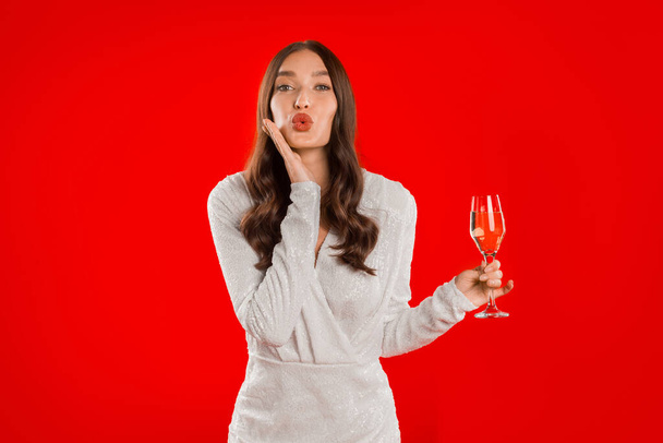 Весела елегантна біла жінка в сукні дме поцілунок на камеру, з келихом шампанського в руці на червоному студійному фоні. Святкування свята, концепція вечірнього банера - Фото, зображення