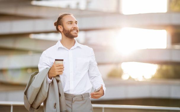 Красивый молодой бизнесмен со смартфоном и кофе на вынос в руках позирует на открытом воздухе, улыбается тысячелетний предприниматель мужчина перерыв на работе, расслабляясь на улице, глядя в сторону копировального пространства, - Фото, изображение