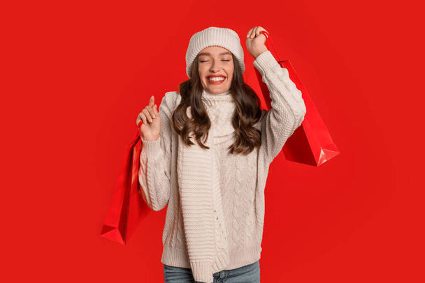 Zimowe oferty wakacyjne. Szczęśliwa pani kupujący trzyma torby na zakupy pozowanie na czerwonym tle studio, świętując początek sezonowej wyprzedaży świątecznej. Baner z radosną zakupową damą trzymającą zakupy - Zdjęcie, obraz