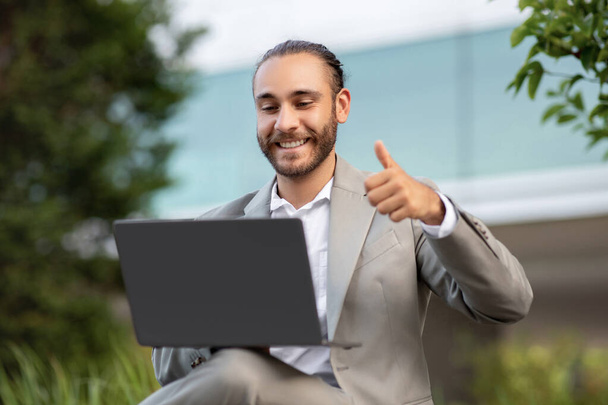Handsome Young Businessman Using Laptop Outdoors, Making Video Call And Showing Thumb Up At Web Camera, Smiling Millennial Male Entrepreneur In Suit Enjoying Online Communication, Closeup - Фото, изображение