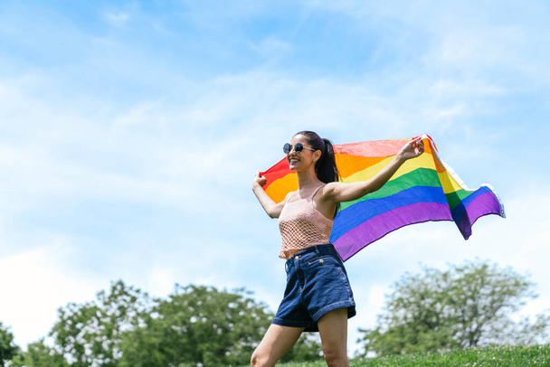 Happy young woman waves LGBT pride flag in park on sunny day, exprimant liberté et joie. - Photo, image