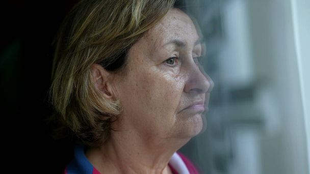 Traurige, in sich gekehrte Seniorin steht am Fenster ihres Hauses und blickt mit besorgter Rührung auf den Blick, Großaufnahme des besorgten älteren Menschen aus Kaukasien - Foto, Bild