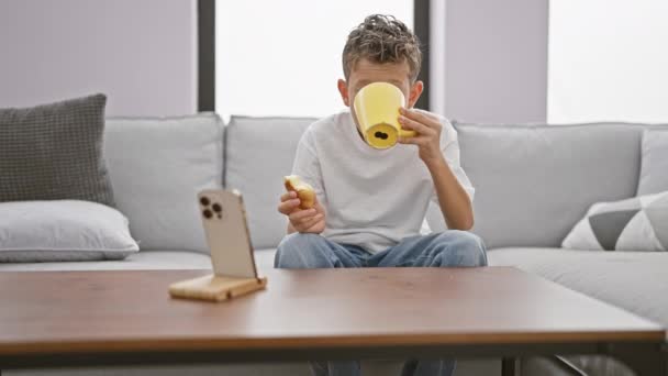 Suloinen pieni vaalea poika, joka katselee videota älypuhelimella, nauttii aamiaiskakusta, joka istuu sohvalla kotona olohuoneessa. - Materiaali, video