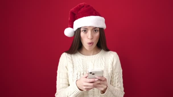Genç, güzel, İspanyol bir kadın yılbaşı şapkası takıyor ve izole edilmiş kırmızı arka plan üzerinde akıllı telefon kullanıyor. - Video, Çekim